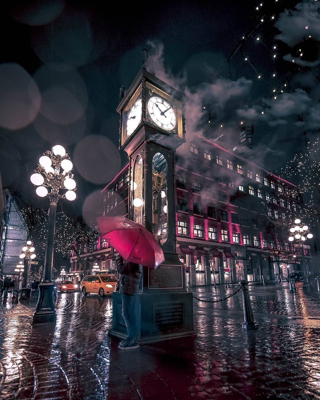 A lone figure with a magenta umbrella beneath the illuminated Gastown steam clock on a rainy Vancouver night